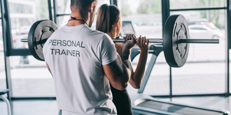 male personal trainer helping sportswoman to do exercises with barbell at gym